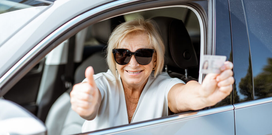An older woman sits happily in the driver's seat holding her reinstated license after it was suspended for impaired driving